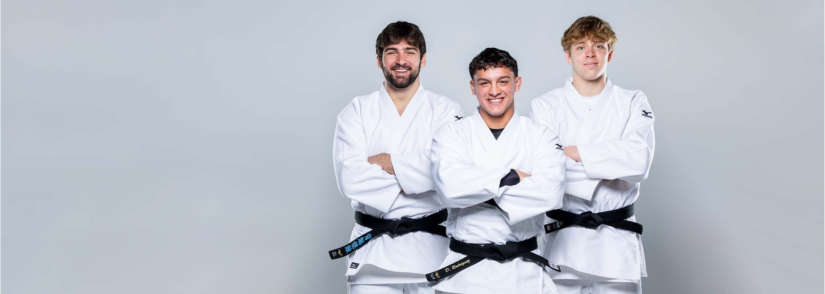 Three young men wearing white judo uniforms and black belts stand side by side with arms crossed, smiling at the camera against a plain light gray background.