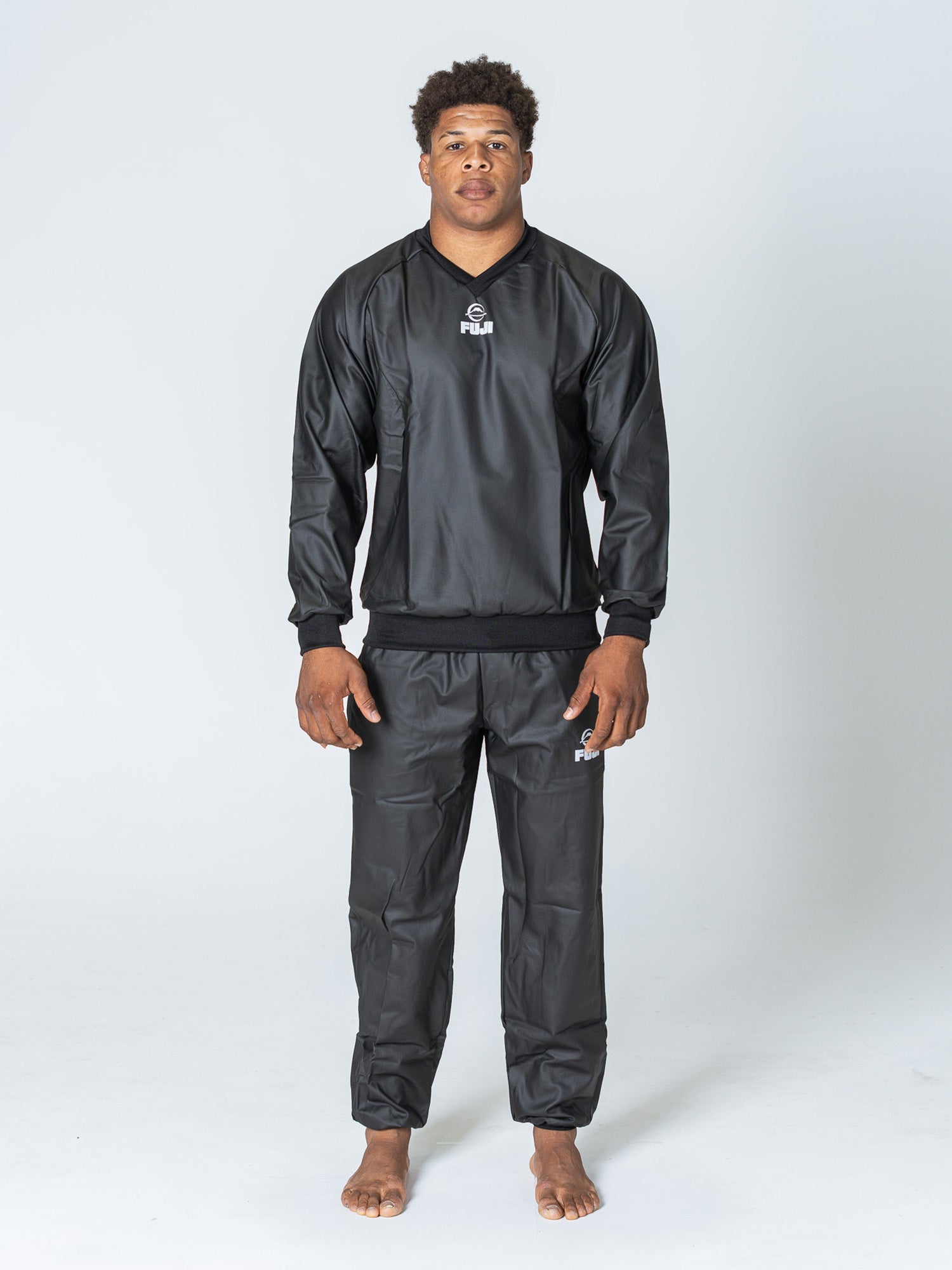 A person stands barefoot on a plain white background, wearing the Grappling Sauna Suit Black—a matching black long-sleeve top and pants set with the Fiji Rugby logo visible on both. They face forward with a neutral expression.