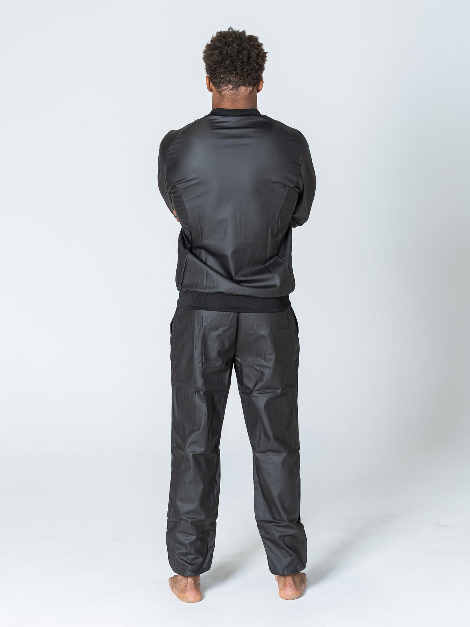 A barefoot person faces away from the camera, arms crossed, wearing the Grappling Sauna Suit Black against a plain white background.