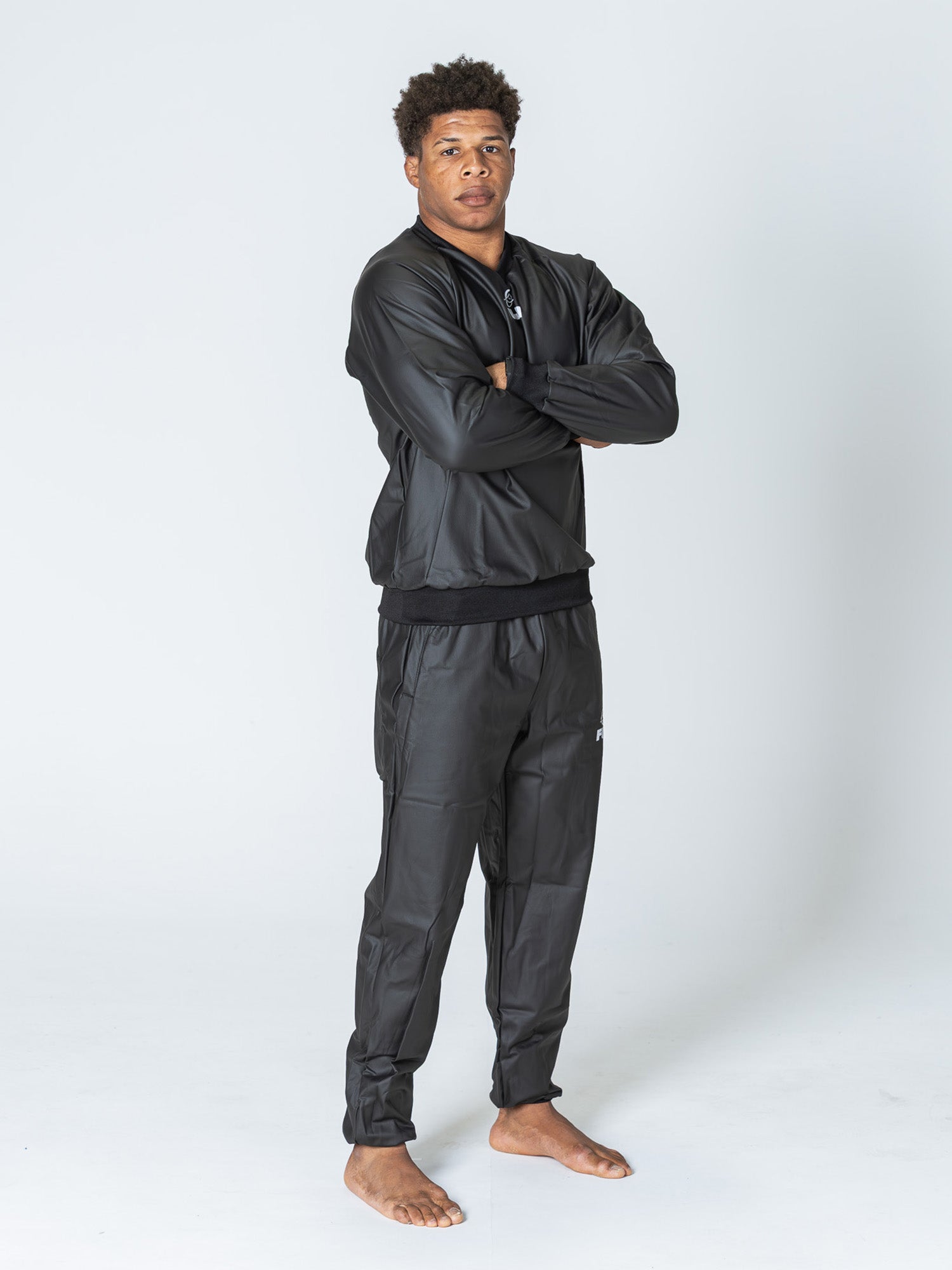A man stands barefoot on a white background with his arms crossed, wearing the Grappling Sauna Suit Black, which features long sleeves and pants.