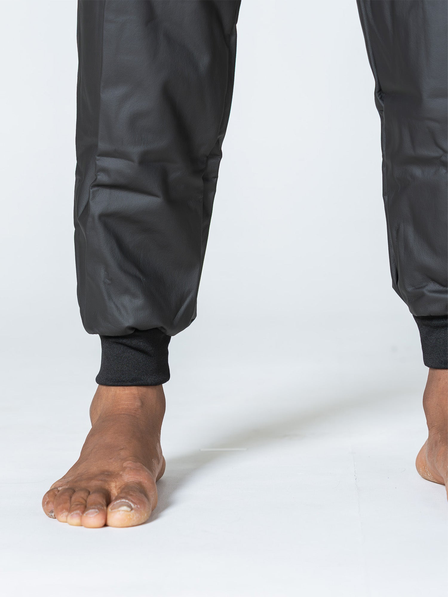 A person wearing the Grappling Sauna Suit Black, standing barefoot on a white surface. The image is cropped to display only the lower legs and feet with fitted cuffs visible.