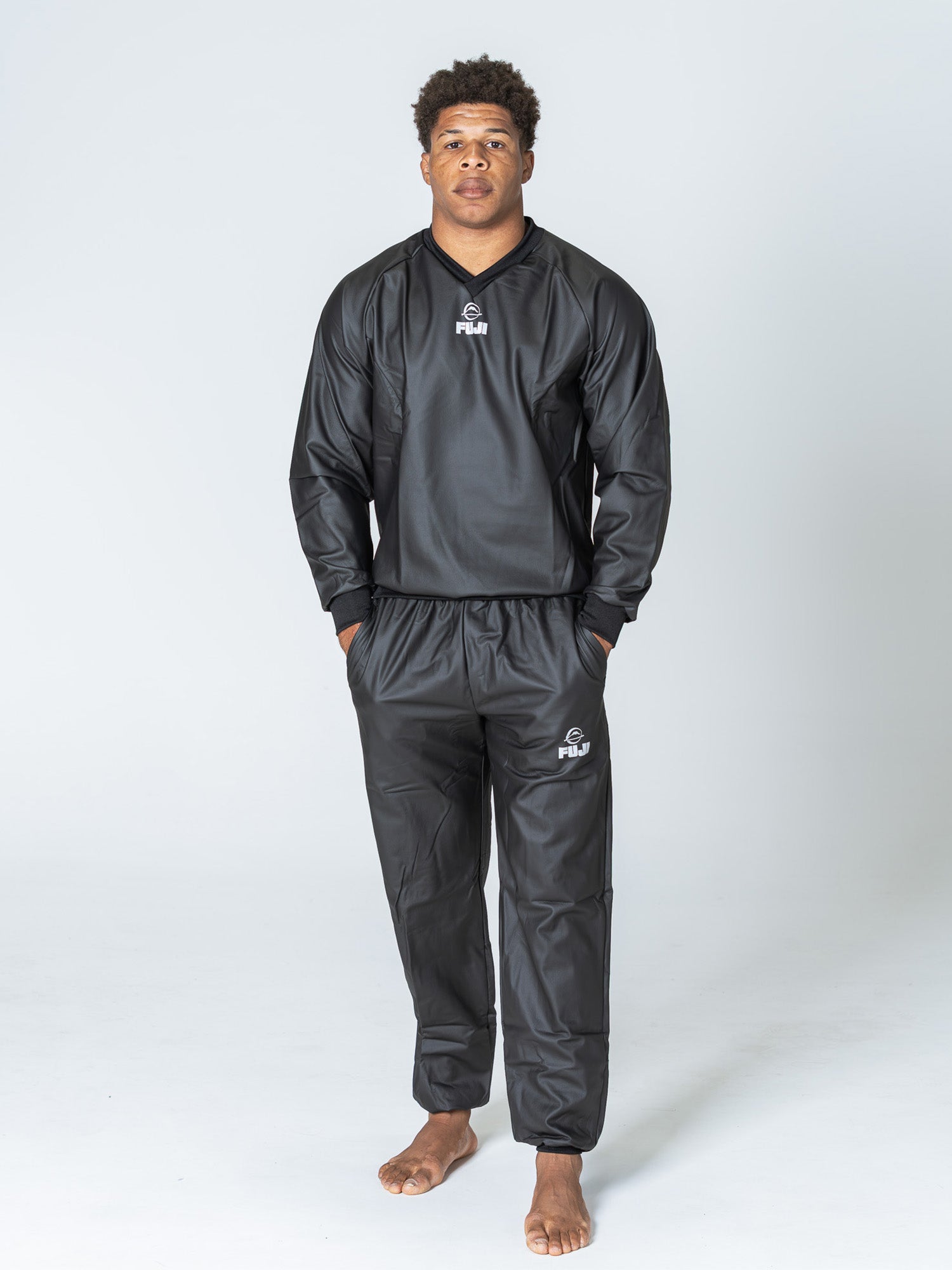A man stands barefoot on a plain white background, wearing the black Grappling Sauna Suit. He faces forward with a neutral expression and his hands in his pockets.