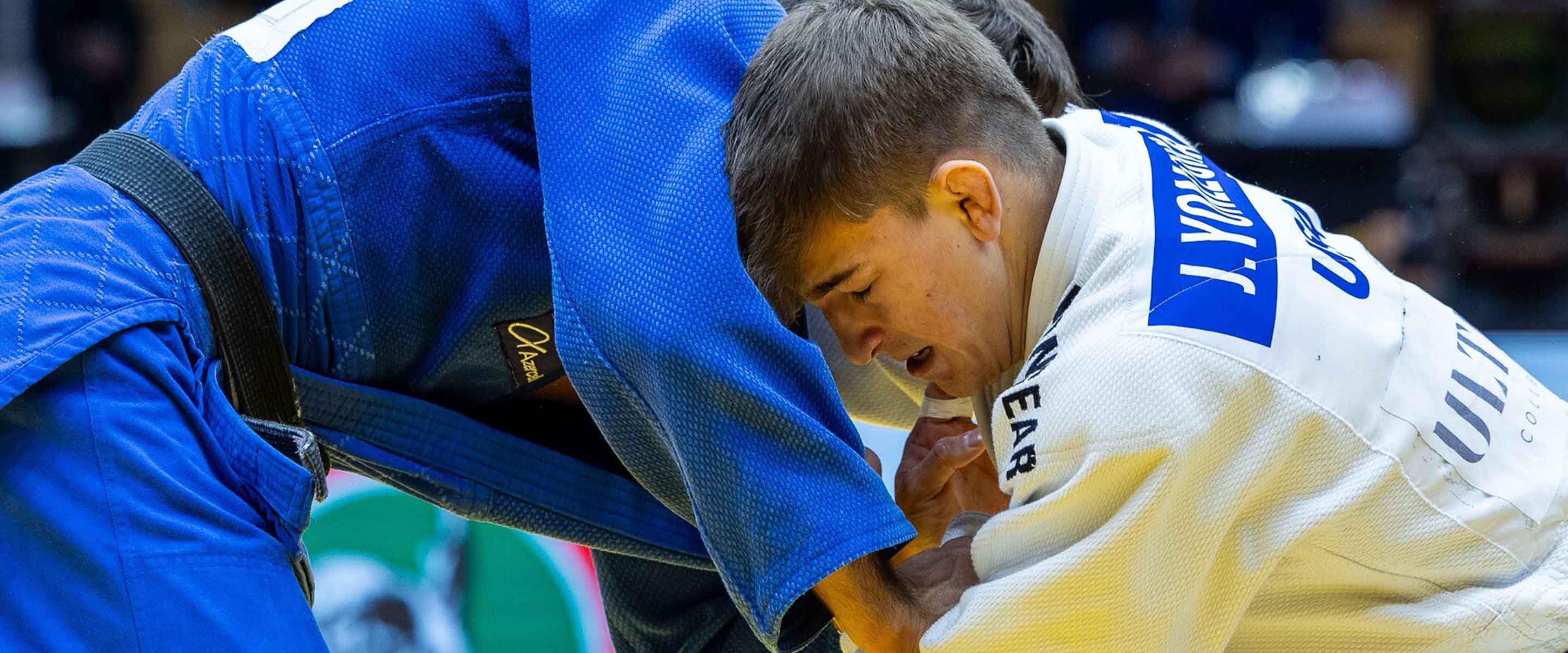 Two judo athletes in blue and white uniforms grapple intensely during a match, focusing on their grips and positioning, with concentration visible on their faces.