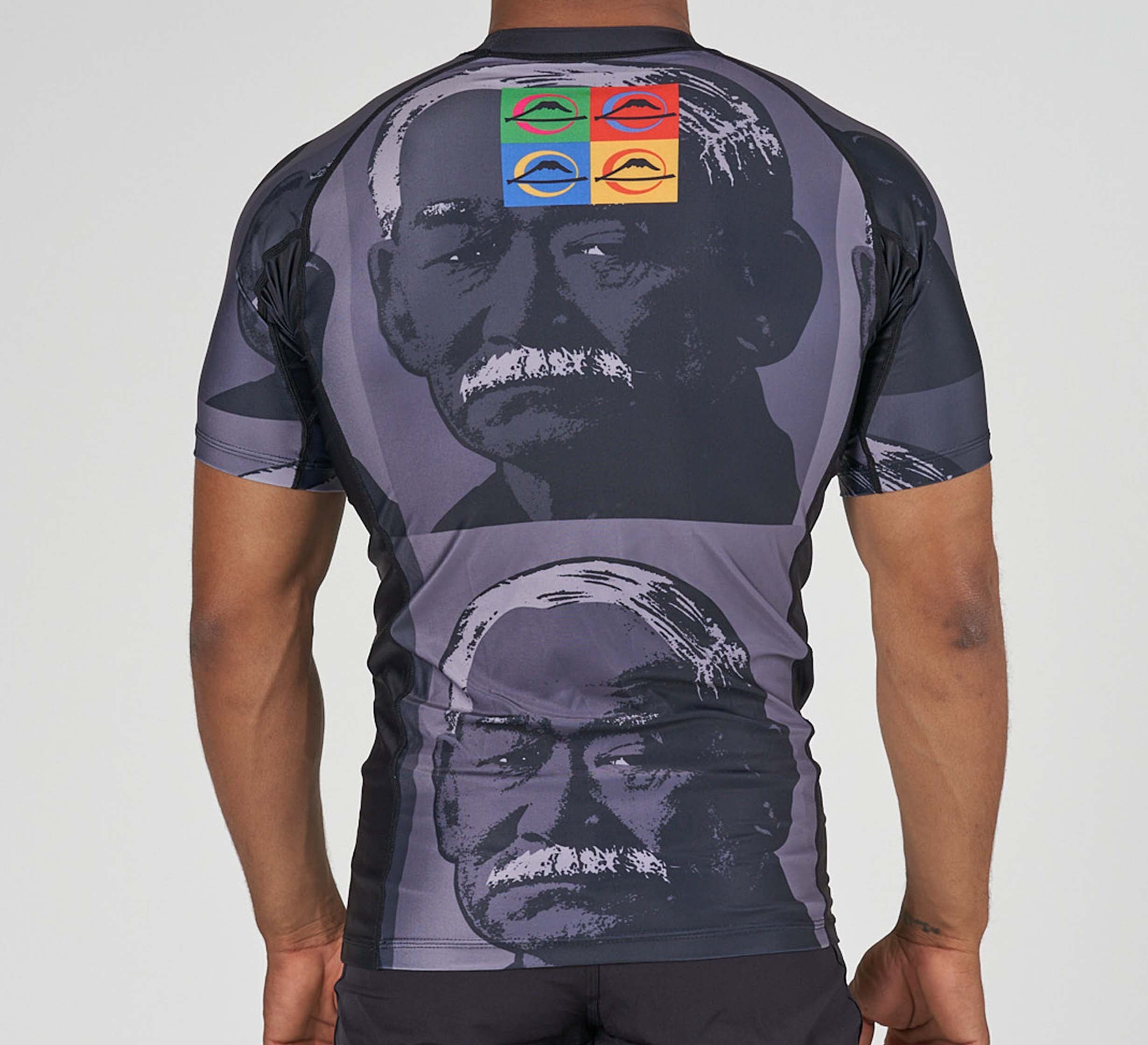 A person wearing the Kano Flex Lite Rashguard, which features a grayscale portrait of an older man with a mustache repeated twice, and four colorful squares above with stylized eyes in red, yellow, green, and blue.