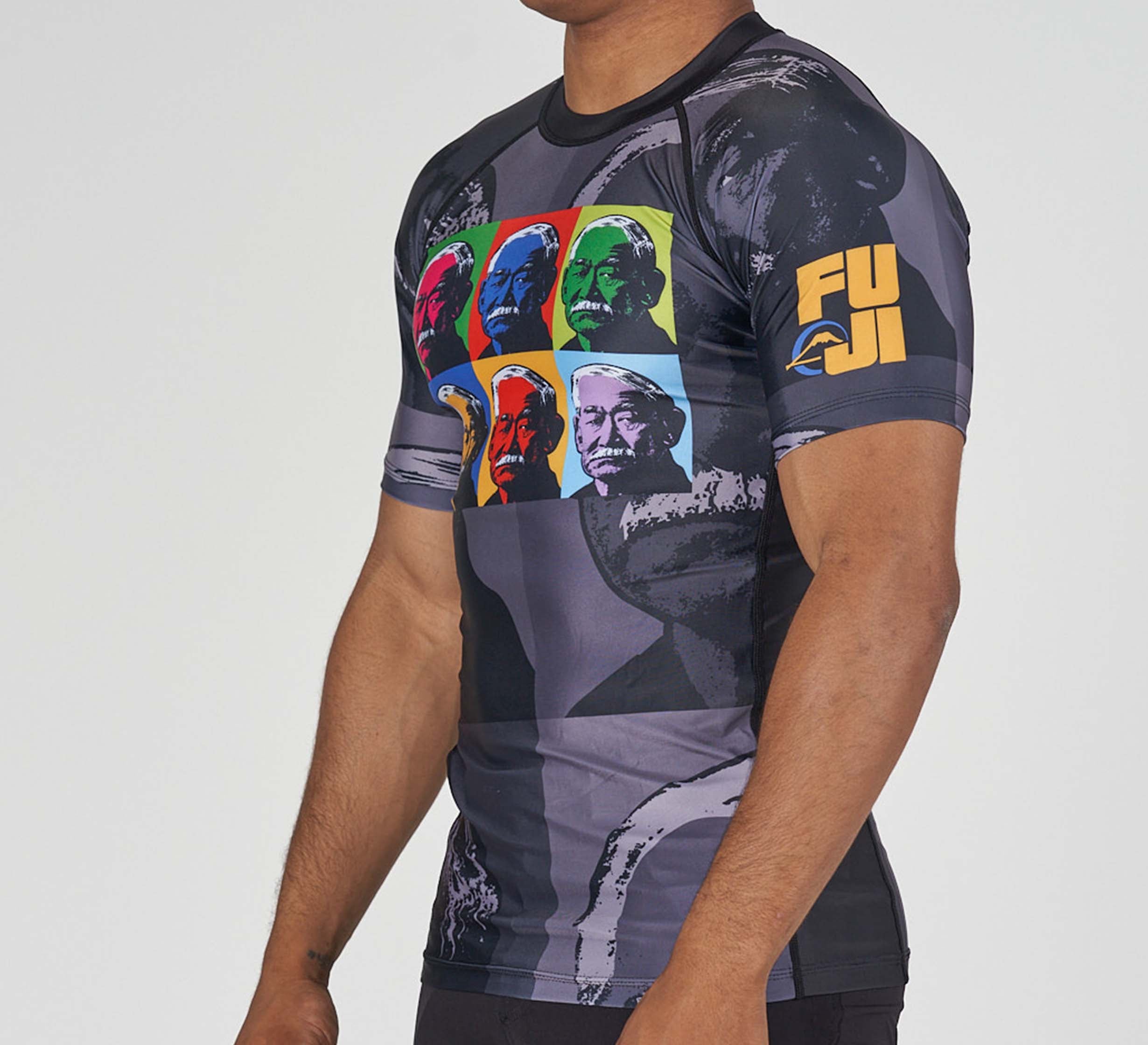 A person wears the Kano Flex Lite Rashguard, featuring vibrant pop art portraits of a man on the front, "FUJI" and a yellow mountain logo on the sleeve, set against a gray background with abstract patterns.