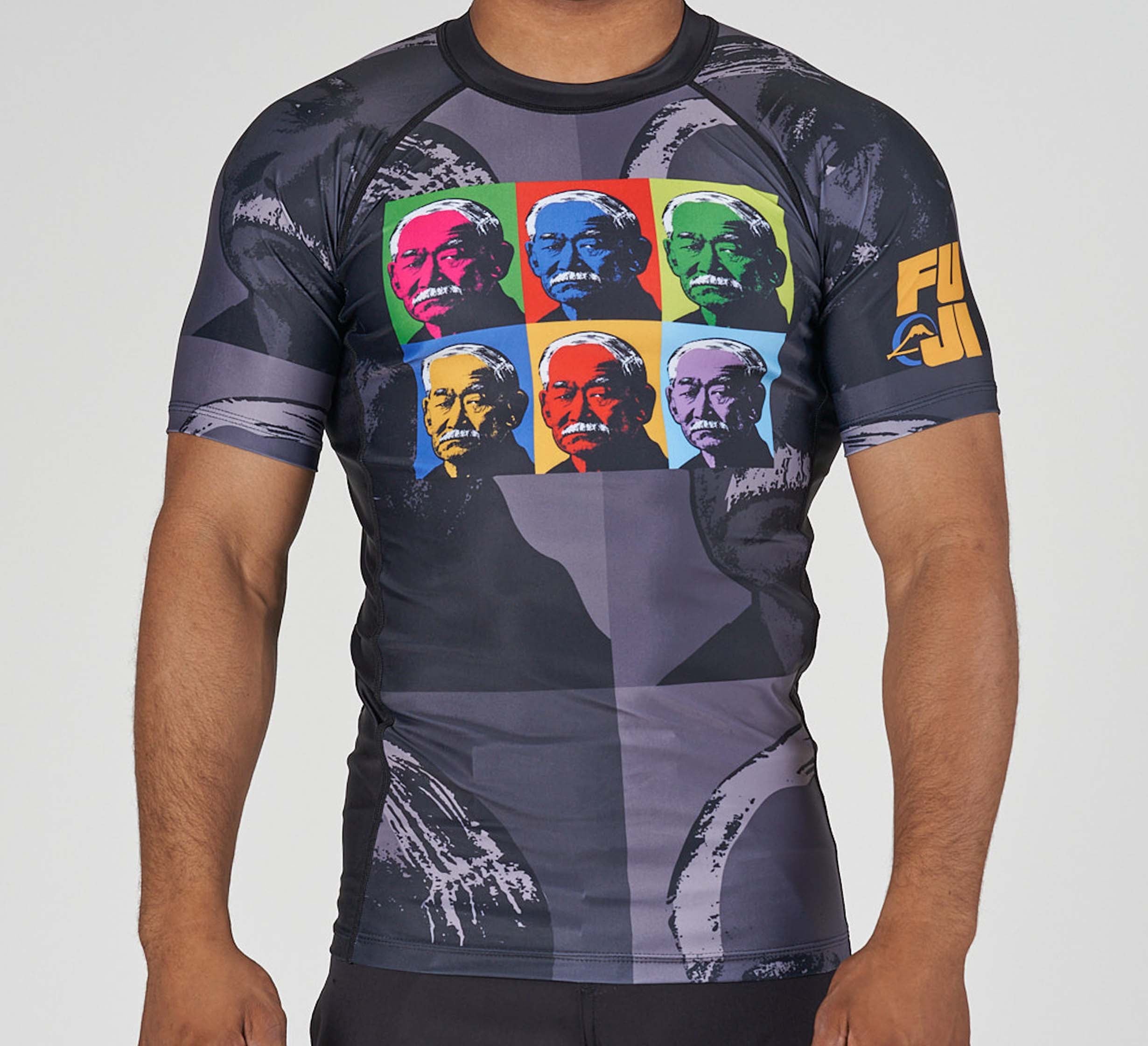 A person wears a Kano Flex Lite Rashguard—short-sleeved, black and gray, with six colorful pop-art portraits of the same older man on the chest and “FUJI” in yellow on the left sleeve.