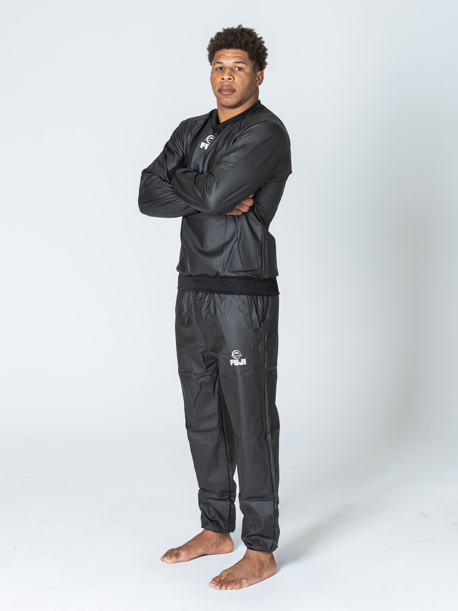 A man stands barefoot on a white background, wearing the Grappling Sauna Suit Black with FUJI logos on the top and pants. His arms are crossed, and he looks at the camera with a neutral expression.