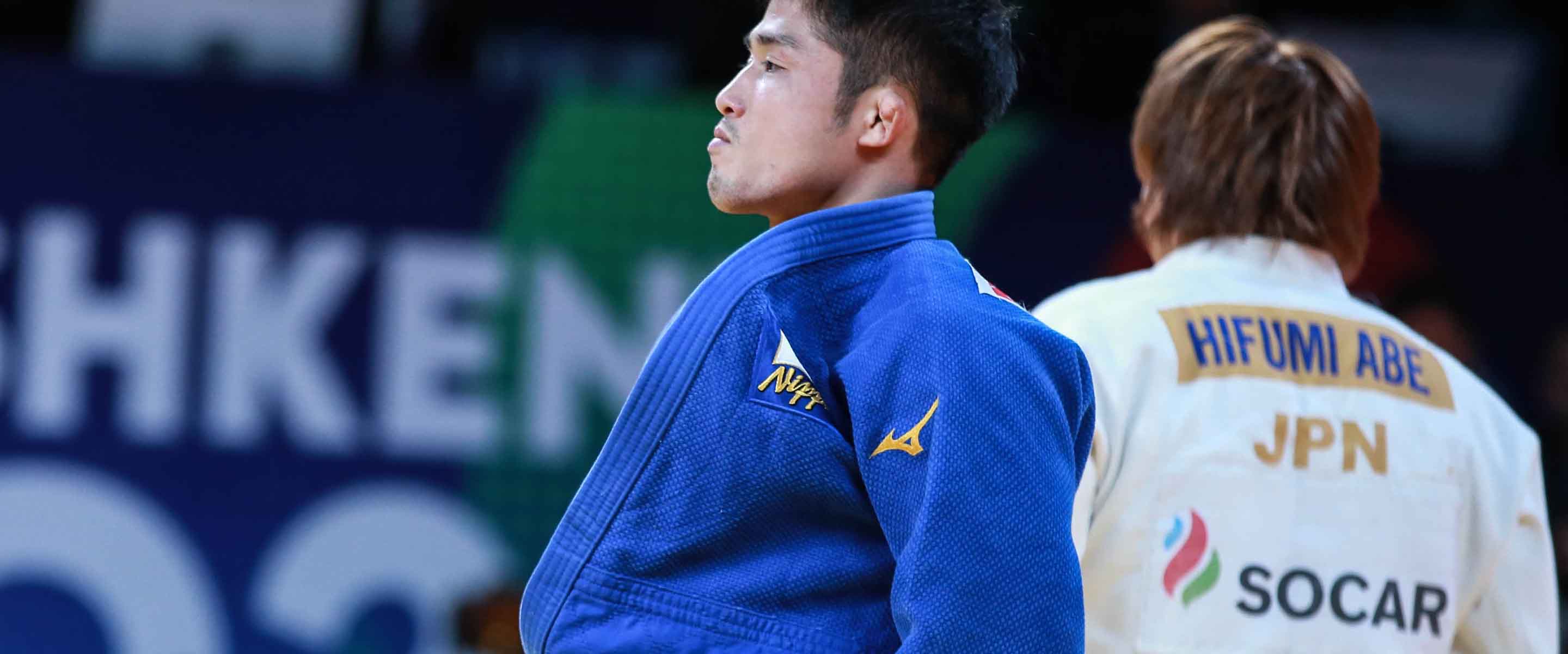A male judo athlete in a blue gi stands with a focused expression, while another athlete in a white gi with Hifumi Abe JPN on the back stands in the background.