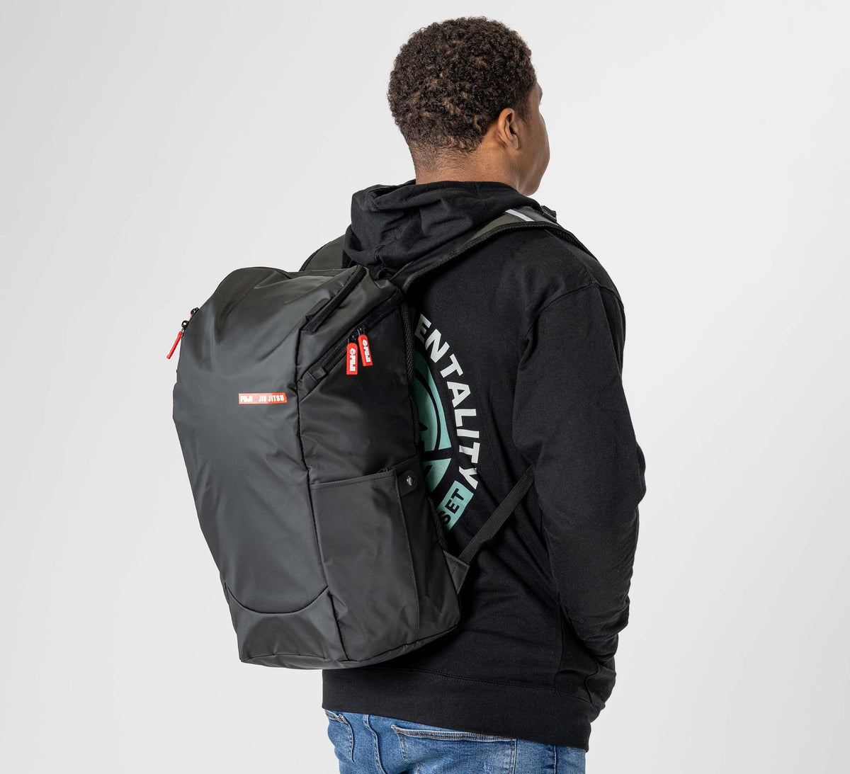 A person in a black hoodie and blue jeans is seen from behind, carrying the Urban Day Backpack Black with red and white labels on one strap and the main pocket, set against a plain light-colored background.